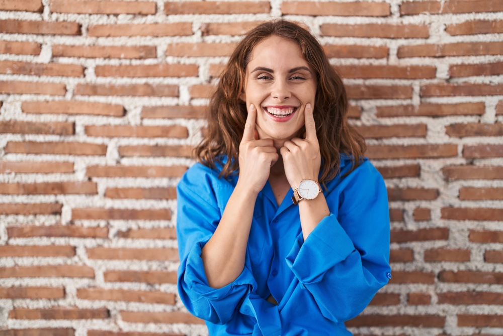 Beautiful brunette woman standing over bricks wall smiling with open mouth, fingers pointing and forcing cheerful smile 