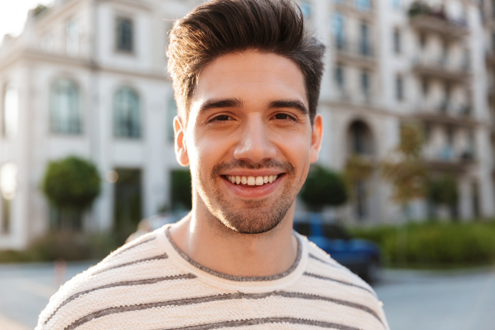 Image of pleased handsome man smiling and looking at camera while walking at city street