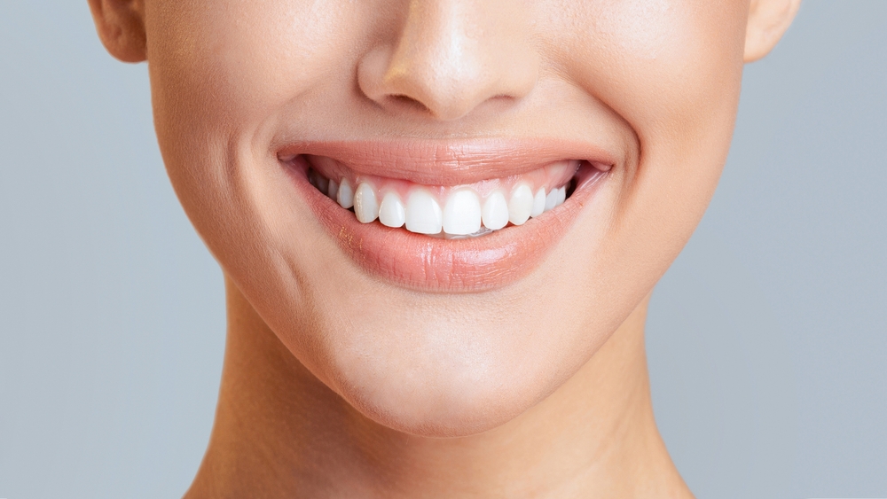 Woman mouth smiling. Her teeth are white and healthy, and she has a gentle smile on her face. The image is likely taken indoors, as the background is a plain blue, cropped